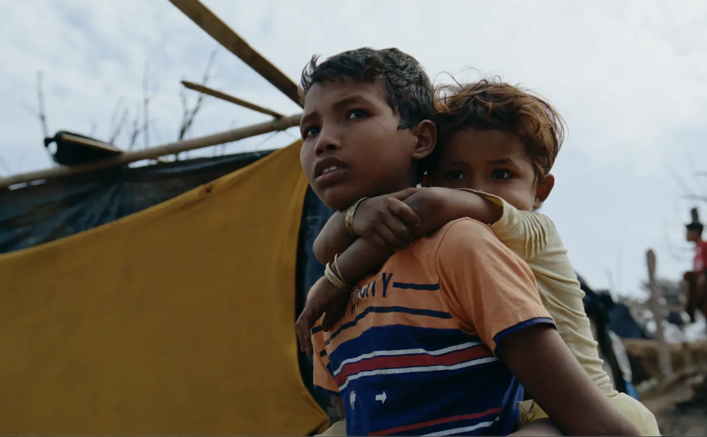 An older child carrying a younger child beside temporary shelters